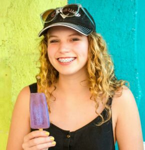 Young girl with hat, glasses, and braces eating a popsicle outside